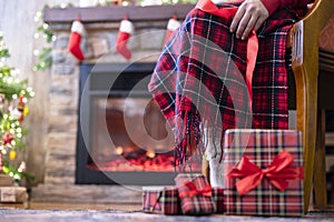 Legs in a socks of disabled women sitting on armchair covered plaid near fireplace and christmas tree  and pakking gift boxes