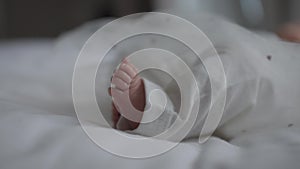 Legs of unrecognizable newborn baby girl in white pants and skirt lying in cozy bedroom on comfortable bed. Close-up