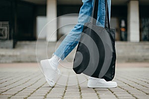 Legs of a woman in jeans and sneakers walking down the street with a black eco bag in her hands, close photo