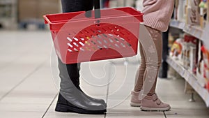 Legs of a woman with a red grocery basket near a shelf in a supermarket. Near a jumping child, trying to get something