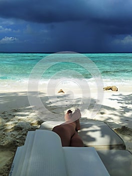 Legs of a woman resting with notepad on a sunbed by the ocean. A beach with the white sand.
