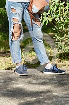 Legs of woman in torn denim trousers and sneakers with bag onbackground of trees in park.