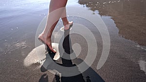 Legs of woman walking barefoot on wet black volcanic sand island beach. Beautiful feet of young girl near sea on sunset