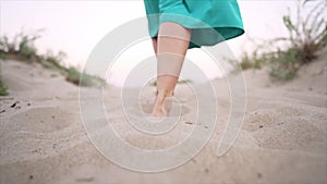 Legs of woman wearing blue skirt walking barefoot on sandy beach or desert. Slow motion. Girl walks away from camera