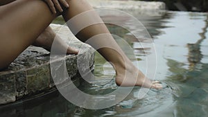 The legs of a young and beautiful European girl in a swimming suit close-up sitting near swimming pool with barefoot wet legs