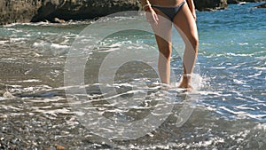Legs of young girl going along sea pebble beach on sunny day. Gentle waves splashing over female feet walking barefoot