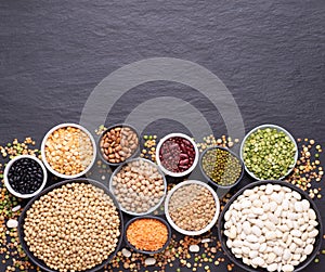 Legumes, lentils, chickpeas and beans assortment in various bowls on black stone background, top view with copy space