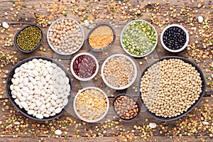 Legumes, lentils, chickpeas and beans assortment in various bowls on wooden background, top view