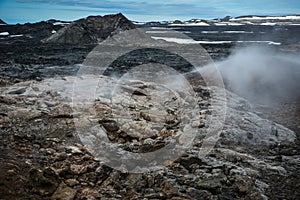 Leirhnjukur old black and red lava field with colorful stones and smoke coming from ground and blue sky in Iceland, overcast day