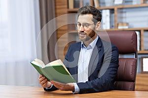 Leisure businessman resting and reading book sitting at desk in business suit inside home office, joyful man improving