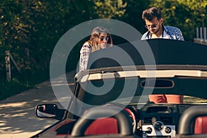 Leisure, road trip, travel and people concept - happy friends pushing broken cabriolet car along country road