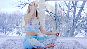 Leisure time, happy girl quenches thirst while relaxing in lotus pose on background of window indoors