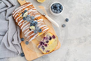 Lemon blueberry cake with lemon icing and fresh berries on top on the board on a gray concrete background with cup of tea.