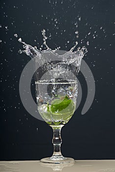 Lemon falling into a glass of transparent water causing splashes upwards. Isolated on gray background