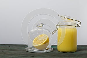 Lemon Kurd in a glass jar. Nearby is half a lemon in a container with a glass lid. Stand on brushed boards, on a white background
