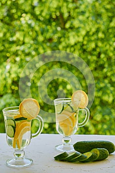 Lemon water in two glasses with ice and cucumber slices on the blurred nature background. Summer cold cocktails. Healthy drinks