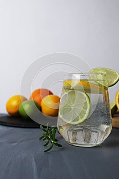 lemonade citrus orange lime lemon in a glass with ice and rosemary on a gray background next to the fruits close-up. Preparation