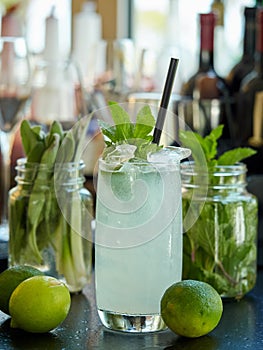 Lemonade drink in a glass with ice and mint leaves with a straw wine bottles on the background on a bar counter , two limes