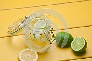 Lemonade with ice, lemon and lime in a jar on a yellow wooden ba