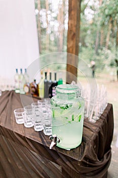 Lemonade jar and glasses for alcohol drinks on wooden table outdoors. Rustic catering at wedding reception. Barn wedding