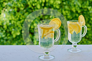 Lemonade water in the glasses on the blurred nature background with copy space. Summer cold cocktails with fresh sliced lemons