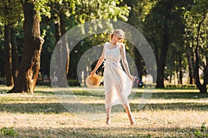Length view of beautiful girl in white dress holding straw hat while smiling and standing on meadow with closed eyes