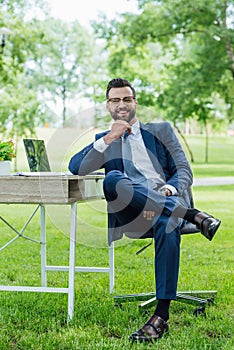 Length view of businessman sitting in office chair near table with laptop and plant, smiling and looking at camera