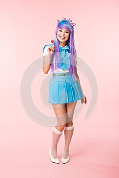 Length view of smiling asian otaku girl holding lollipop on pink