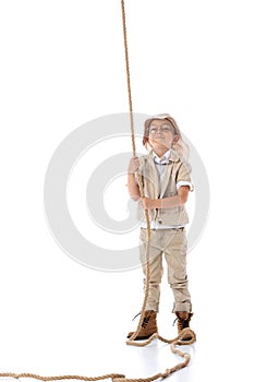 Length view of smiling explorer kid in hat and glasses holding rope on white