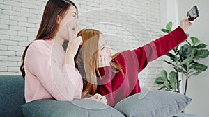 Lesbian Asian couple using smartphone selfie in living room at home, sweet couple enjoy funny moment while lying on the sofa when
