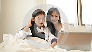 A Lesbian couple is watching a movie from a computer laptop while sitting together on the bed.