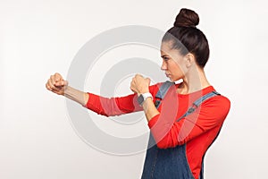 Let`s fight! Side view of determined confident girl with hair bun in denim overalls punching, boxing with clenched fists, struggl