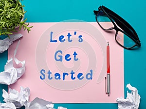 Let's get started message - Office desk table with supplies top view. crumled paper, pen, glasses and flower.