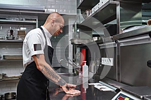 Let`s start. Young bald male chef with beautiful tattoos on his arms looking at order lists on a steel table in a