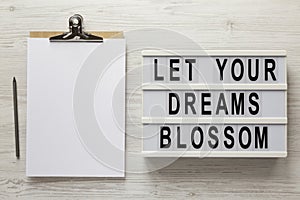 `Let your dreams blossom` on a lightbox, clipboard with blank sheet of paper on a white wooden surface, top view. Flat lay,