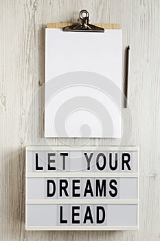 `Let your dreams lead` on a lightbox, clipboard with blank sheet of paper on a white wooden surface, top view. Flat lay, overhea