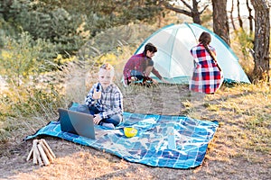 Lets go camping. Family camping. Reach destination place. Two girls pitch tent and one boy watching show on the laptop, sitting on