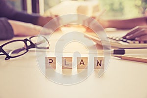 The letter `PLAN` is on the table with business people working behind the scenes - Business team collaboration ideas.