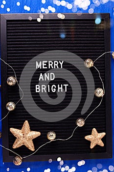 Letterboard with phrase merry and bright and golden decorations  on bright blue paper textured background.