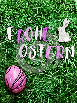 The letters of Happy Easter in German, a pink marbled egg and a decorative little bunny on green easter grass