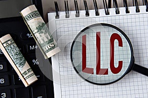 Letters LLC under a magnifying glass on a white notebook against the background of a fragment of a calculator and banknotes