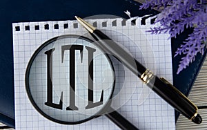 The letters LTL under a magnifying glass on a white piece of paper against the background of a fragment of a notebook and a pen