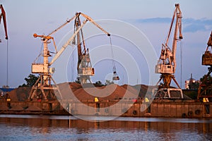 Level-luffing bulk-handling cranes load sand onto a barge in a river port in the evening light