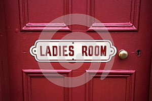 Old vintage metal painted sign saying Ladies Room on a painted red wooden background with brass door handle