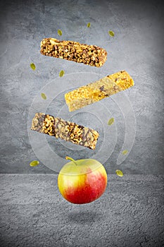 Levitating food: three muesli bars, ripe yellow red apple and a few green seeds over gray surface