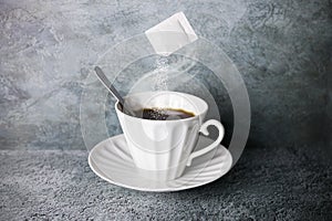 Levitating sachet of pouring sugar over white porcelain hot coffee cup with steam, spoon and saucer on gray surface