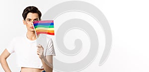 Lgbtq community. Attractive queer man with flitter on face, waving pride rainbow flag and looking at camera, standing in