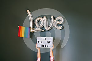 LGBTQ pride month background. Female hands holding lightbox with Text Love is all we need above silver foil balloon with