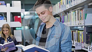 Library. Smiling male student reading book near shelves