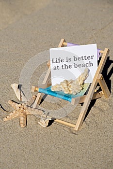 LIFE IS BETTER AT THE BEACH text on paper greeting card on background of beach chair lounge starfish summer vacation